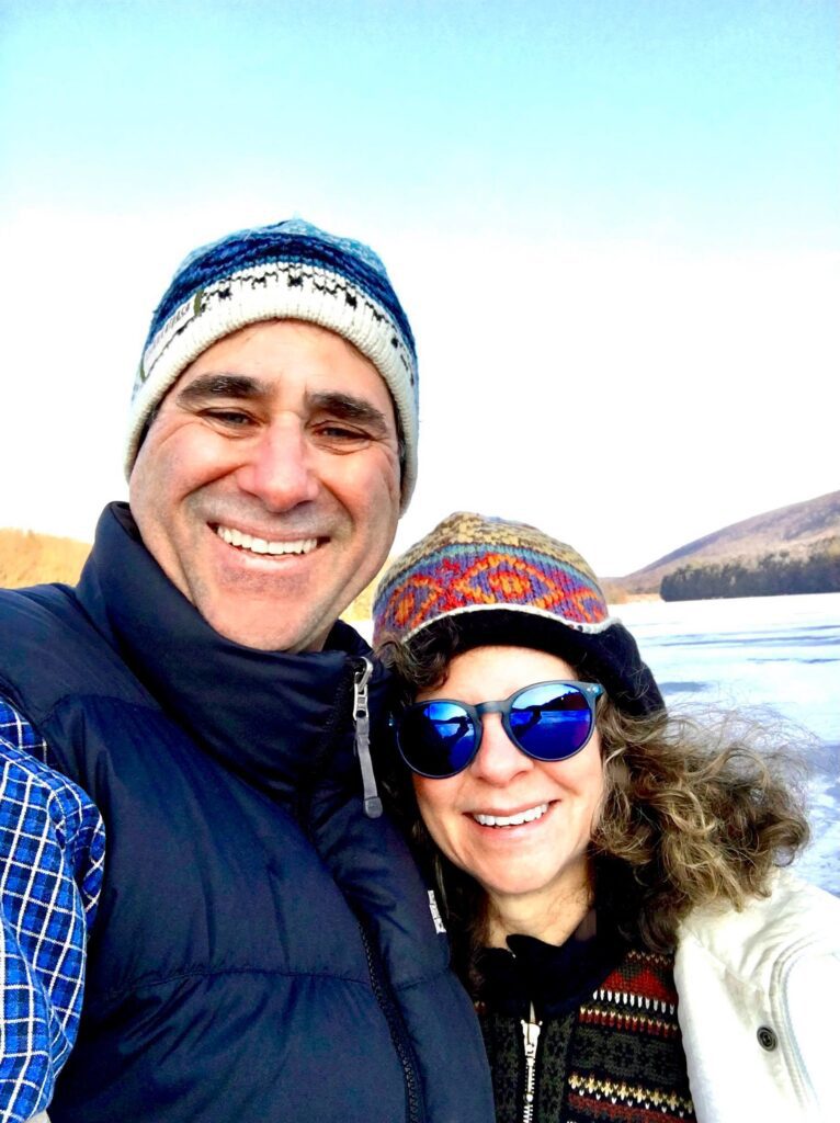 Lynn and Brad on the ice in winter coats and knitted hats, a frozen lake and low hills behind them.